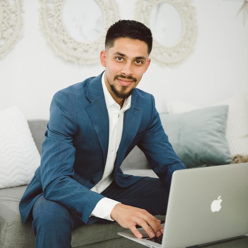 Smiling professional man in a suit working on a laptop in a cozy, stylish home office or living room, surrounded by decorative mirrors and cushions, demonstrating remote work or business success.